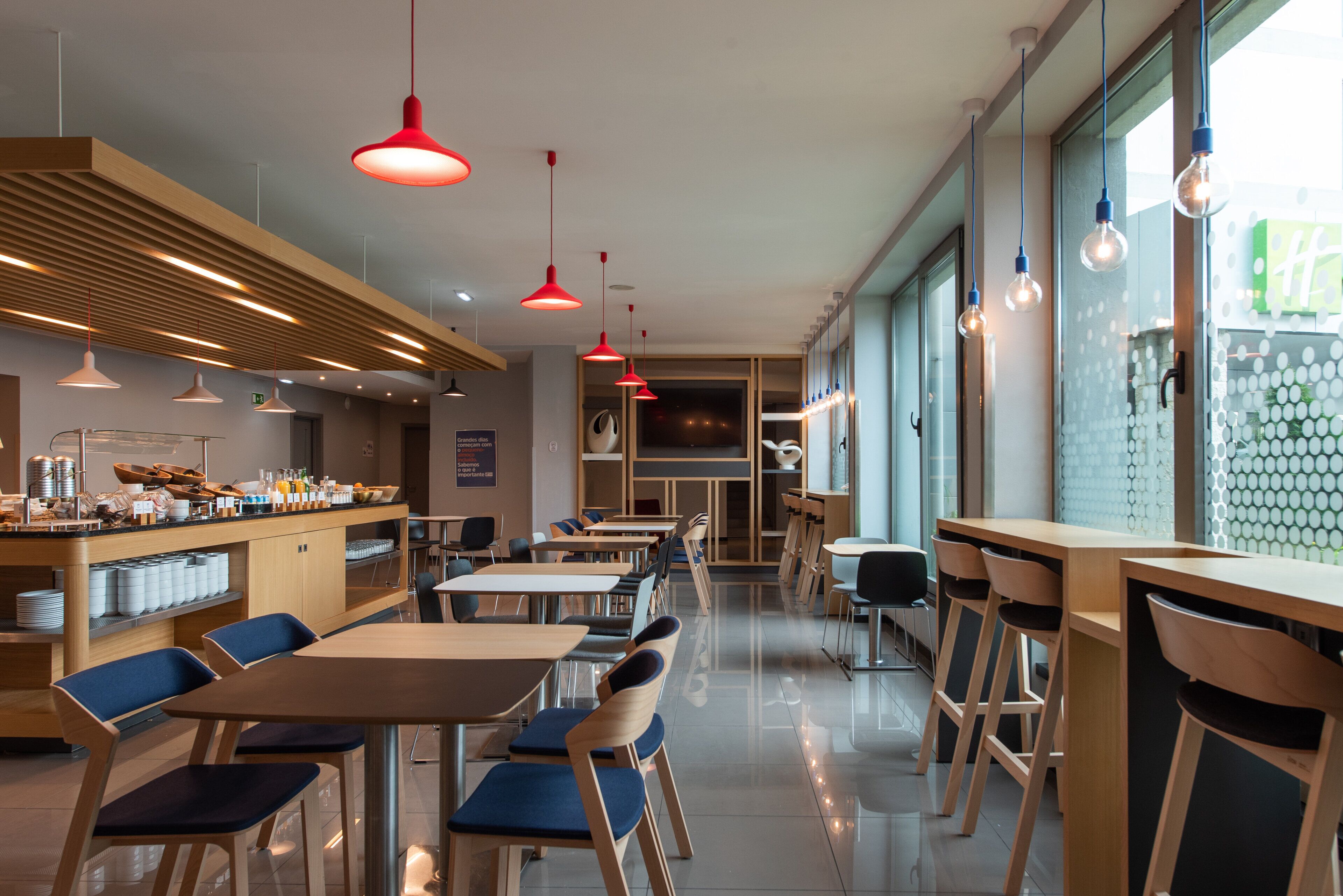 Een modern restaurant met houten tafels en stoelen, rode hanglampen en een balie met etenswaren.