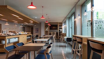Un restaurant moderne avec des tables et des chaises en bois, des lampes suspendues rouges et un comptoir où sont exposés des plats.