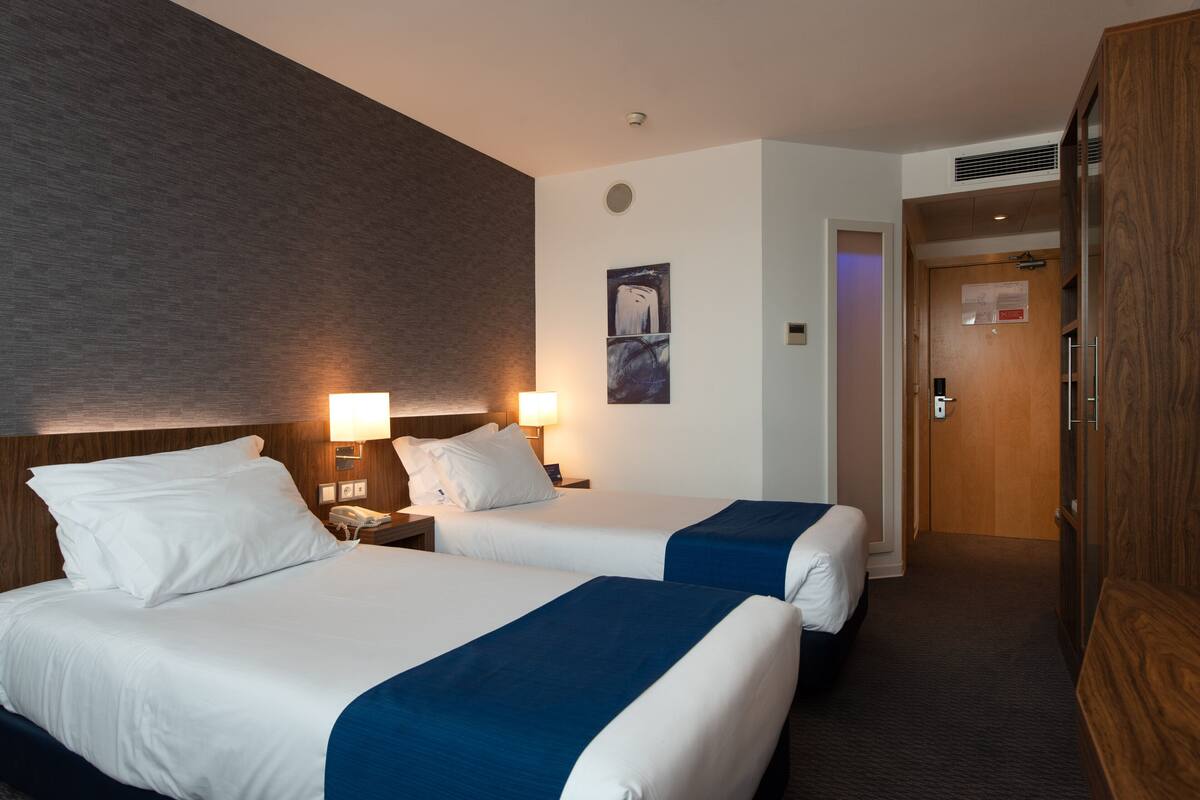 Ett hotellrum med två sängar, en sänggavel i trä, ett nattduksbord med en lampa och en dörr som leder till ett annat rum.