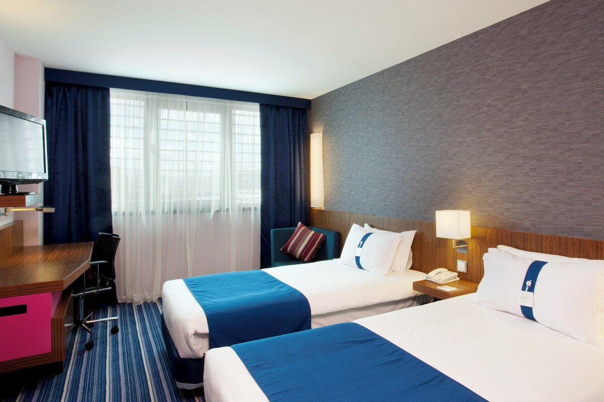 A hotel room with two beds, a desk, a TV, and a window with curtains.
