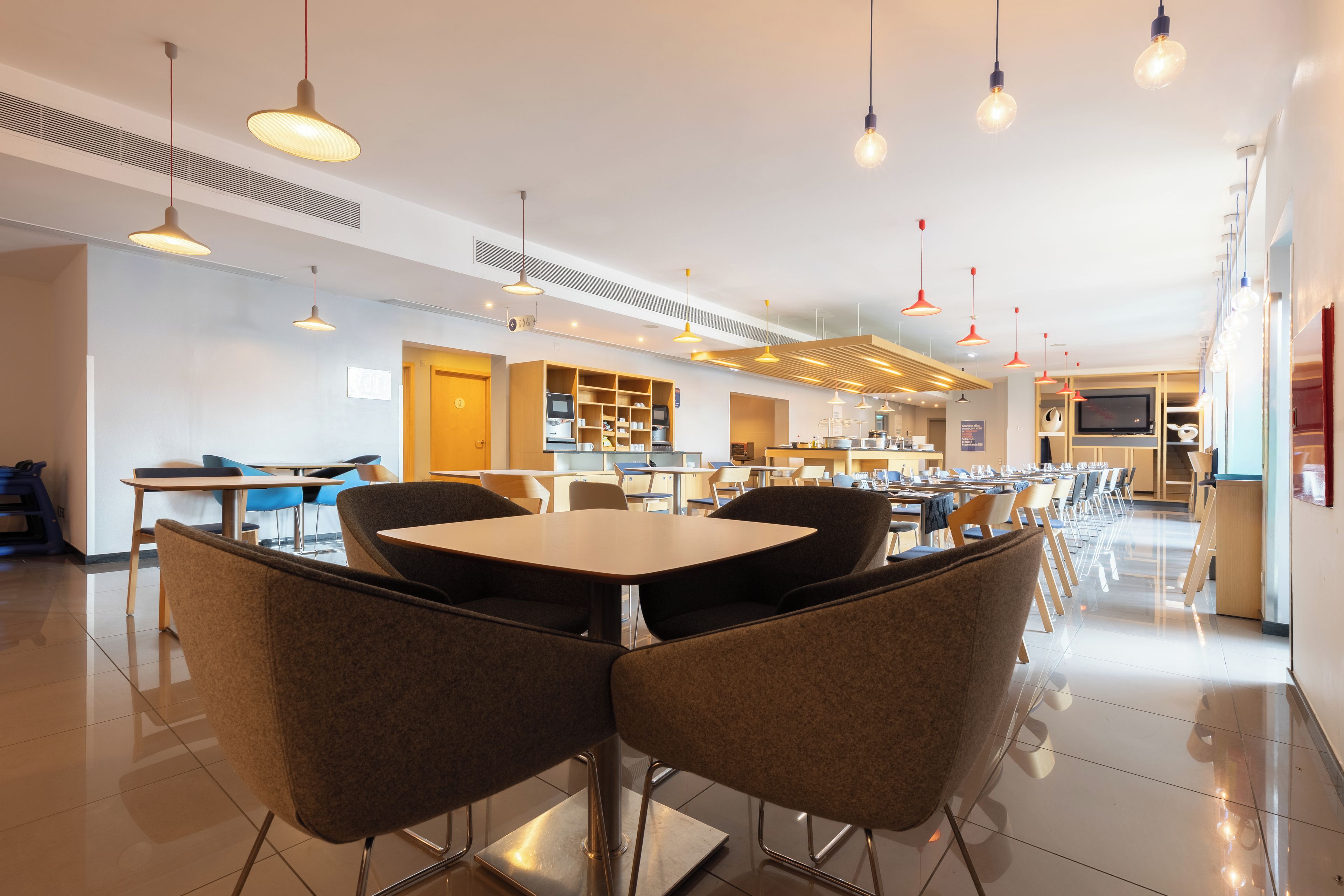 Un comedor moderno con mesas y sillas, una cocina al fondo e iluminación colgante.