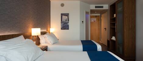Habitación de hotel con dos camas, cabecero de madera, mesita de noche con lámpara y armario.