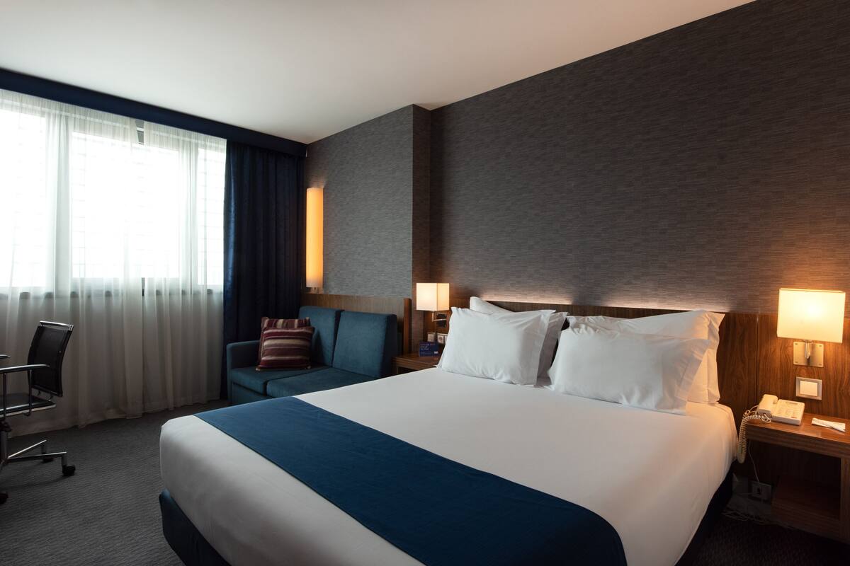 Ett hotellrum med en stor säng, ett skrivbord, en stol och ett fönster med gardiner.