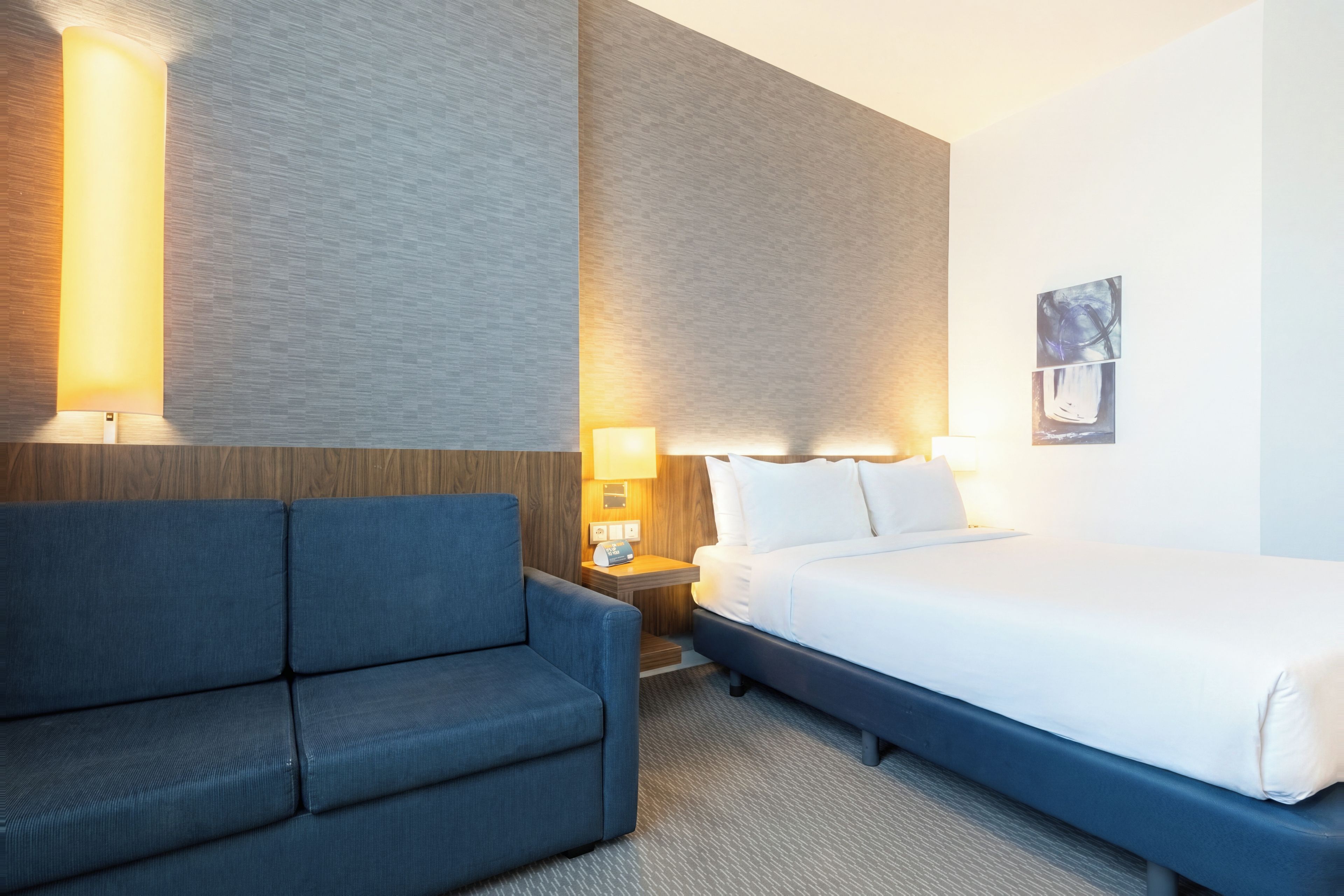 Ett hotellrum med en säng, en soffa, två vägglampor och en tavla på väggen.