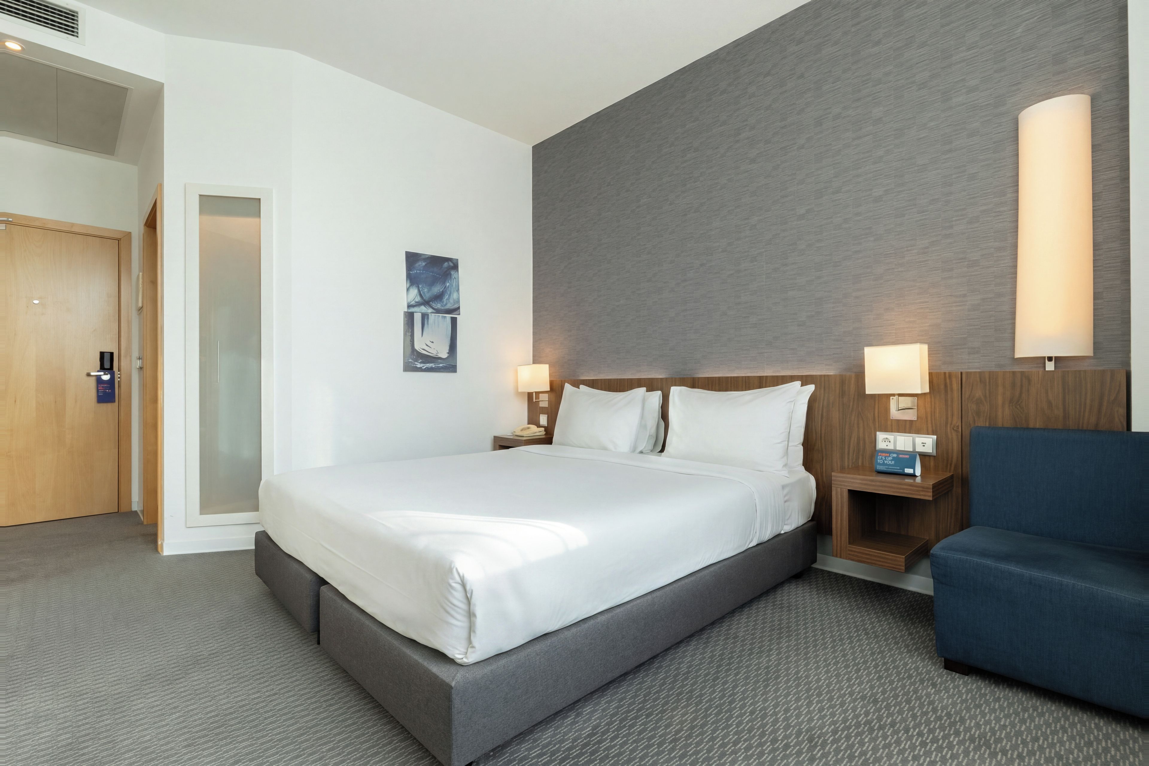 Una habitación de hotel con una cama grande, lámparas de noche, un sillón azul y un cabecero de madera.