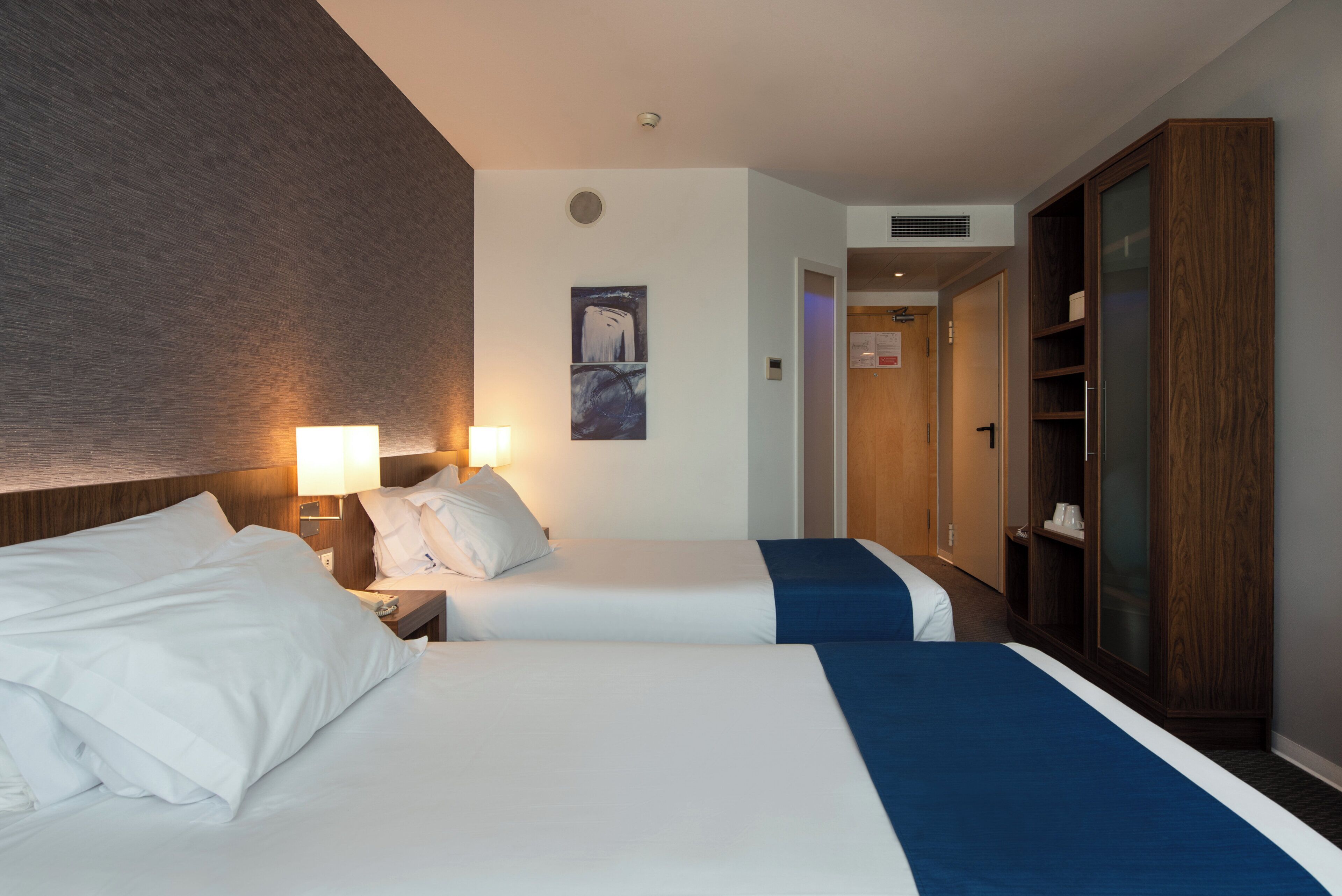 Habitación de hotel con dos camas, un armario, una obra de arte colgada en la pared y una puerta que da a otra habitación.