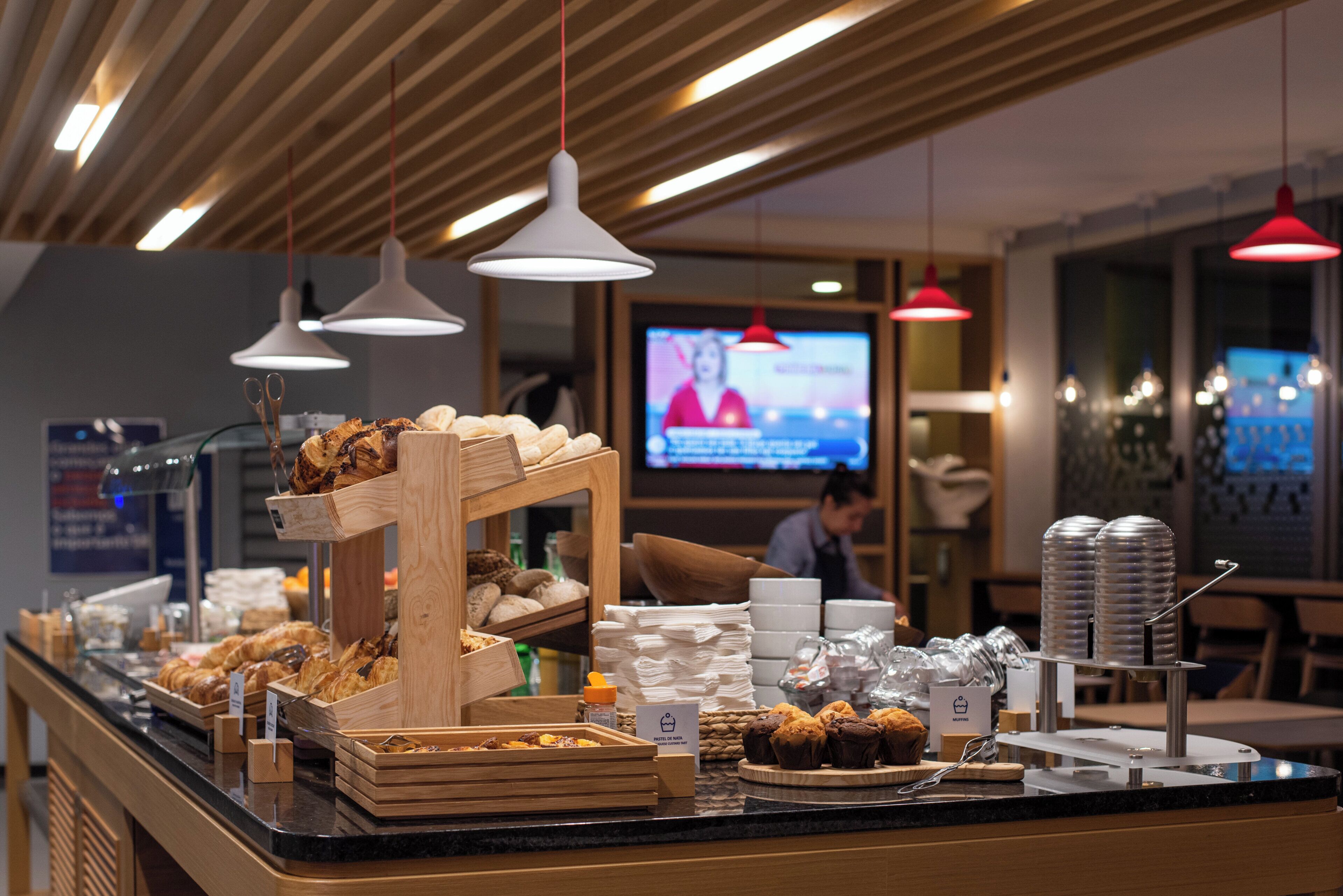A breakfast buffet with various pastries and bread, a coffee station, and a television screen in the background.