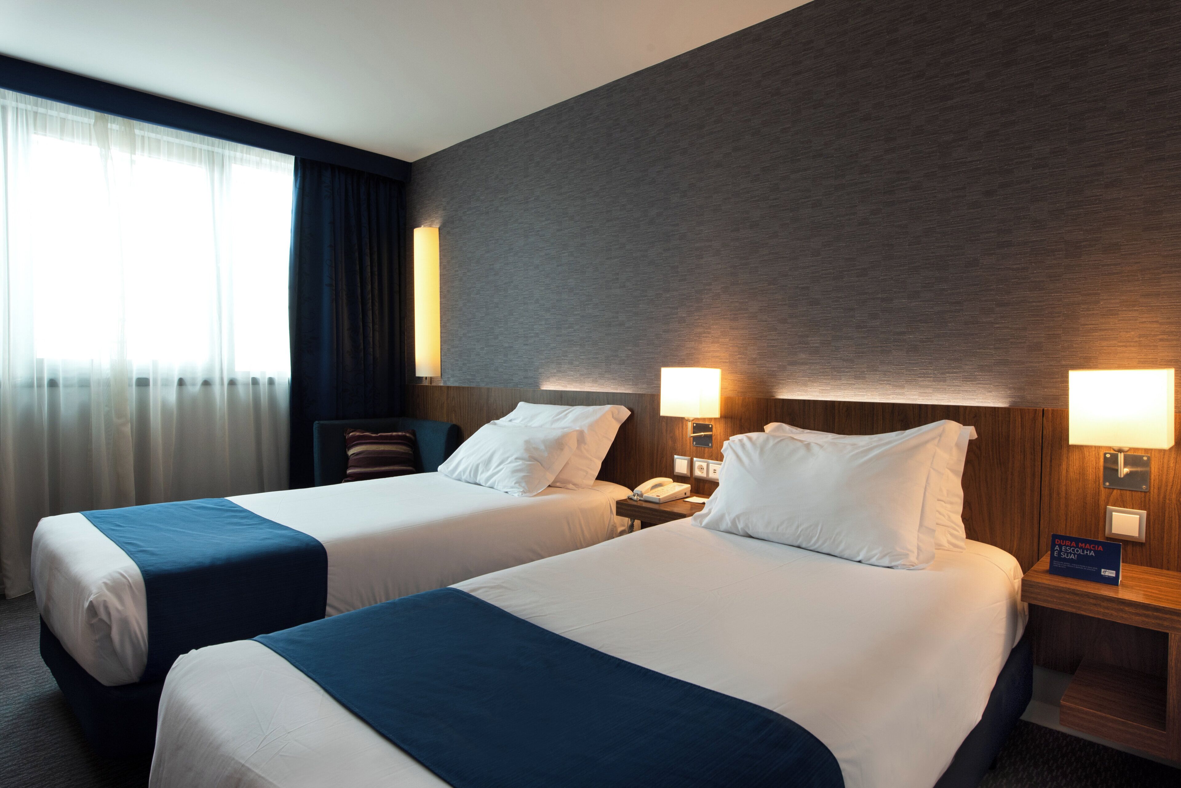 A hotel room with two beds, a nightstand with a lamp, a telephone, and a sign indicating the room number.