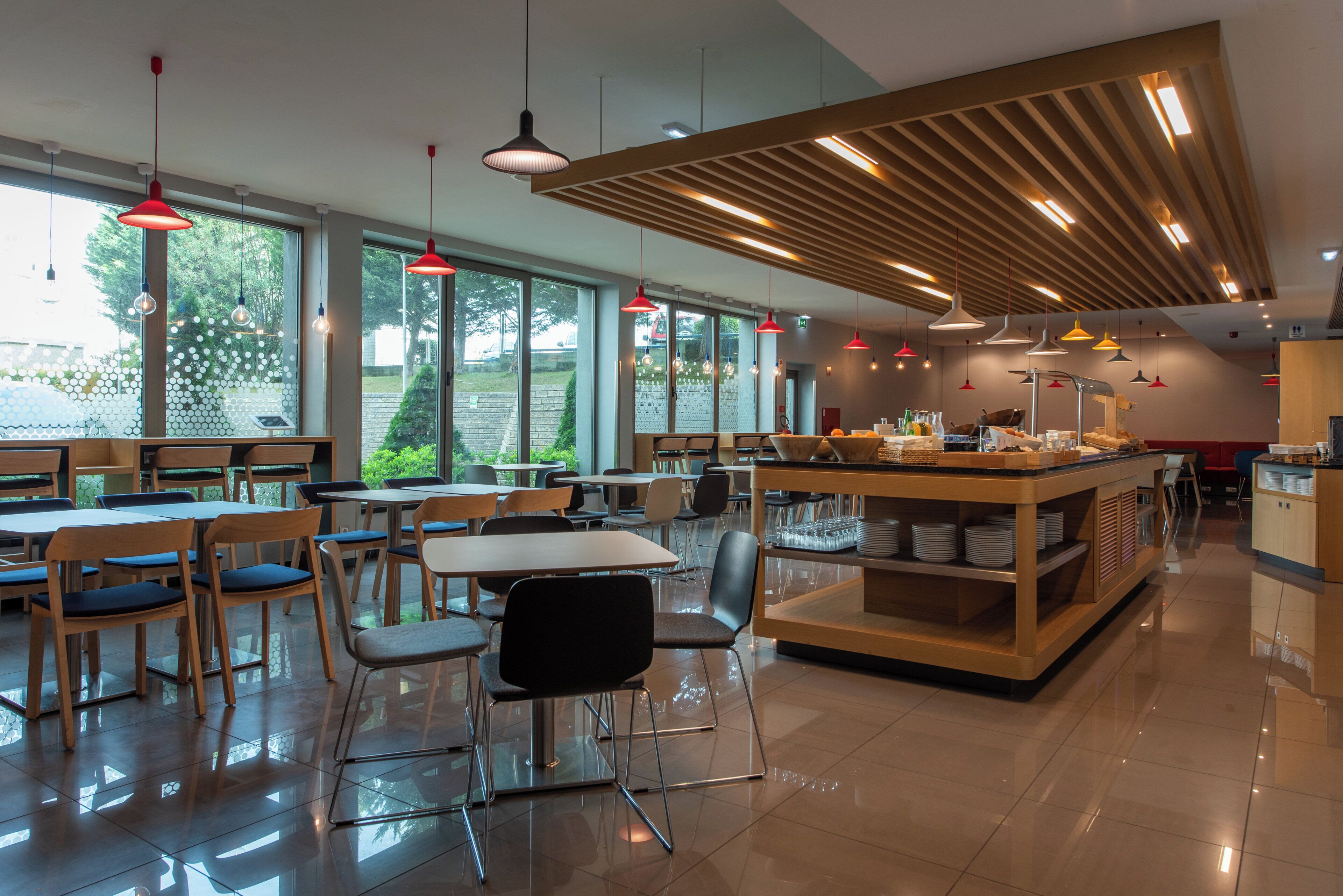 Un interior de cafetería moderno con mesas y sillas de madera, ventanas amplias e iluminación colgante.