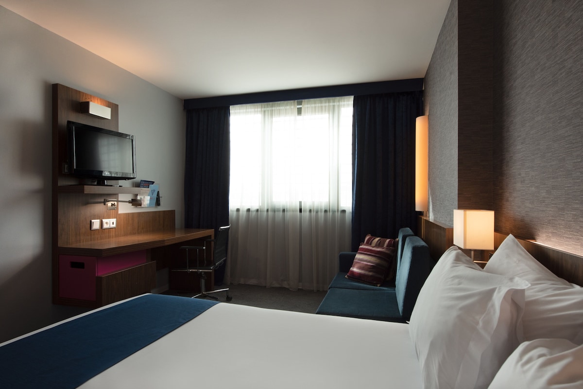 A hotel room with a bed, a desk with a television, a window with curtains, and a wall-mounted lamp.