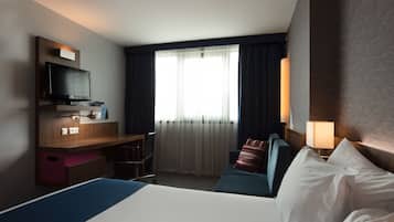 A hotel room with a bed, a desk with a television, a window with curtains, and a wall-mounted lamp.