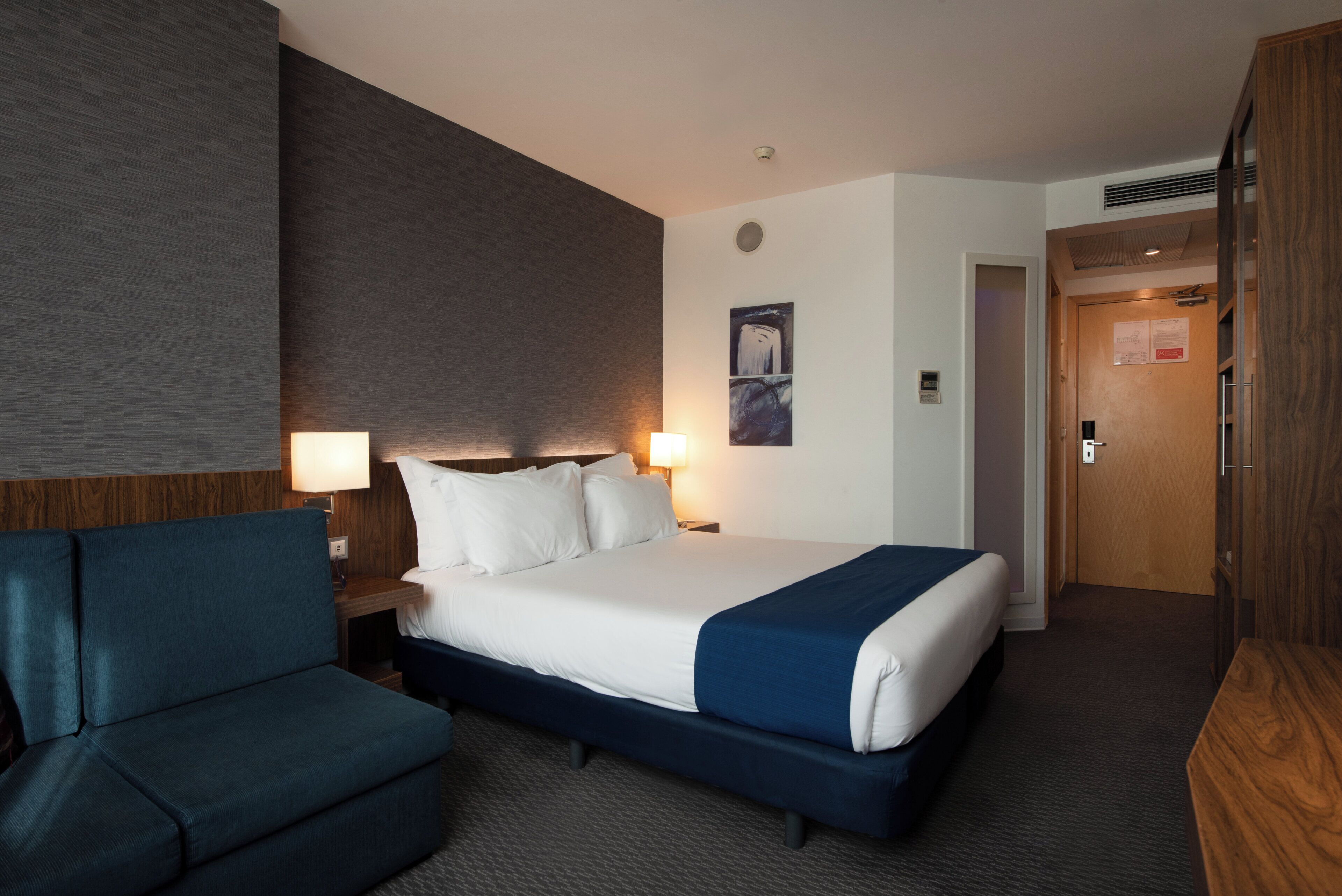 A hotel room with a bed, a desk, a chair, and a door.