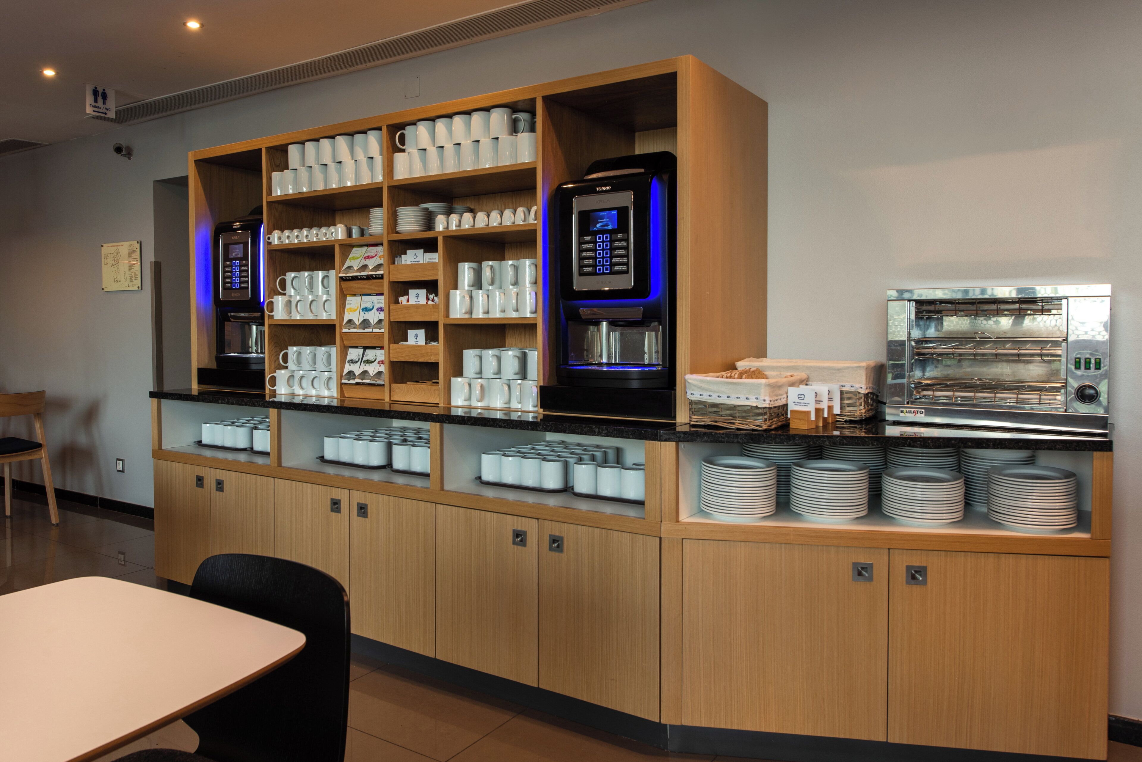 A self-service coffee station with various coffee machines, a food display, and a seating area.