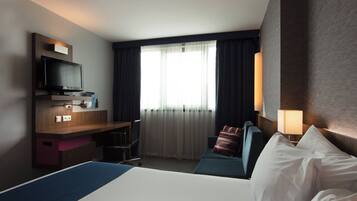 Ein Hotelzimmer mit einem Bett, einem Schreibtisch mit Fernseher, einem Fenster mit Vorhängen und einer Wandlampe.