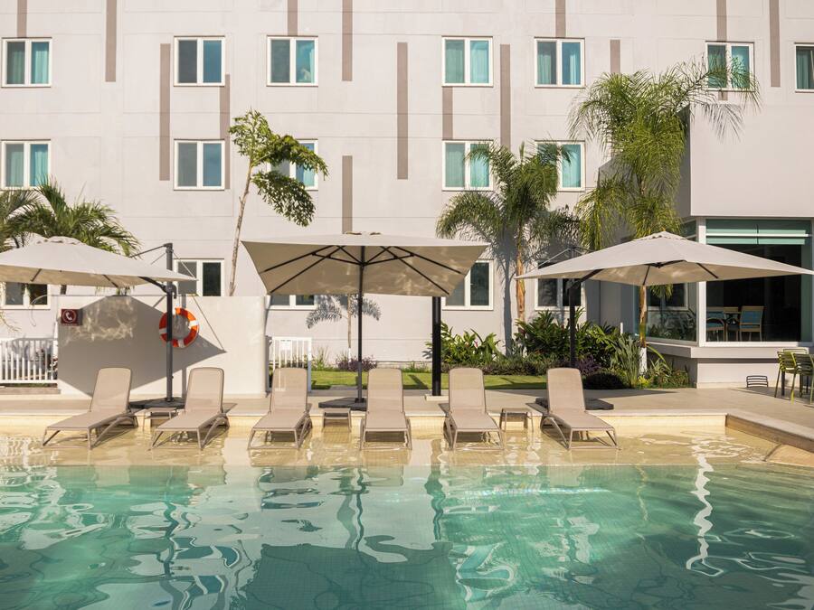A pool area with lounge chairs and umbrellas, surrounded by a building with multiple windows and palm trees.