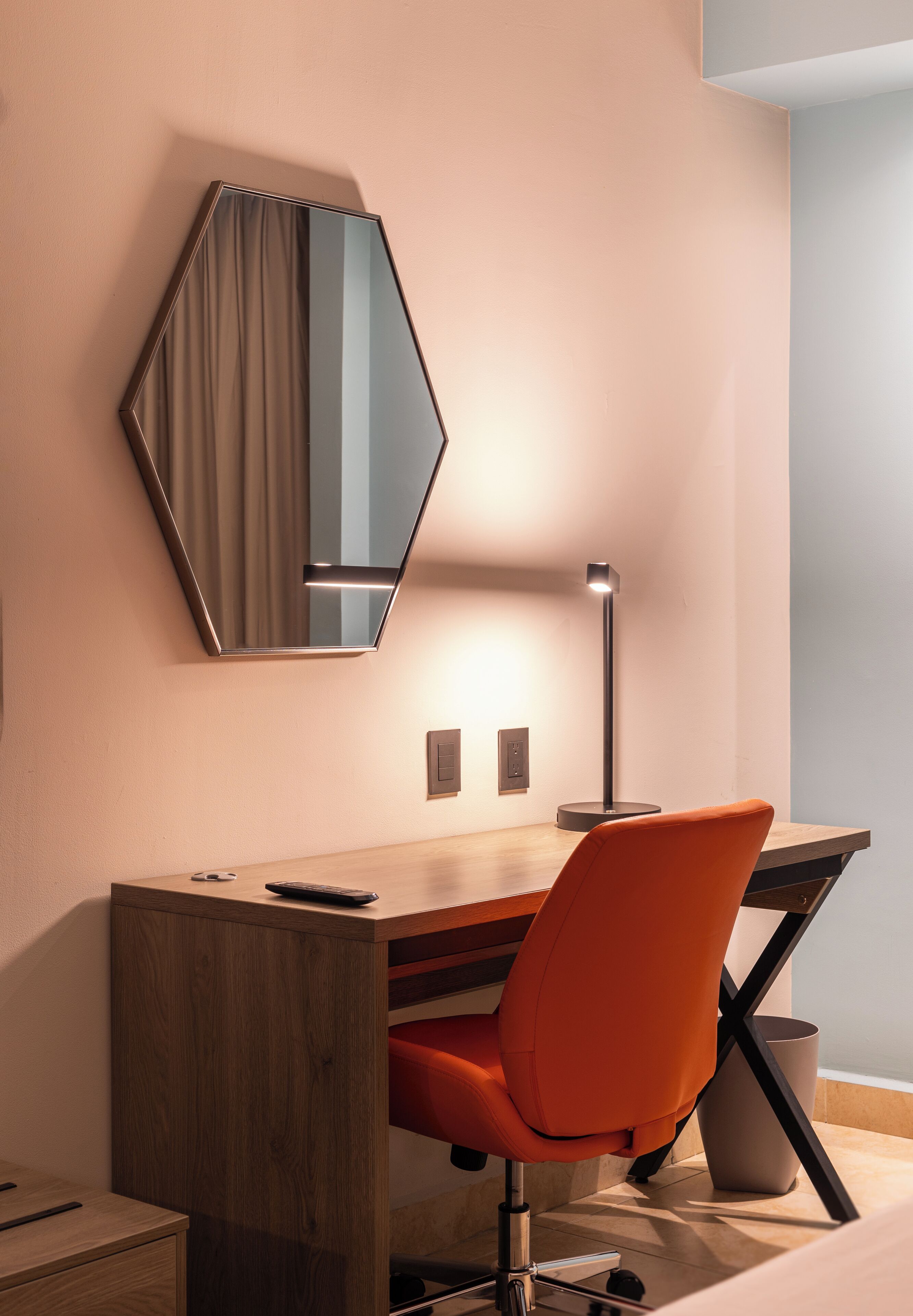 A modern hotel room with a wooden desk, an orange chair, a floor lamp, and a large mirror on the wall.