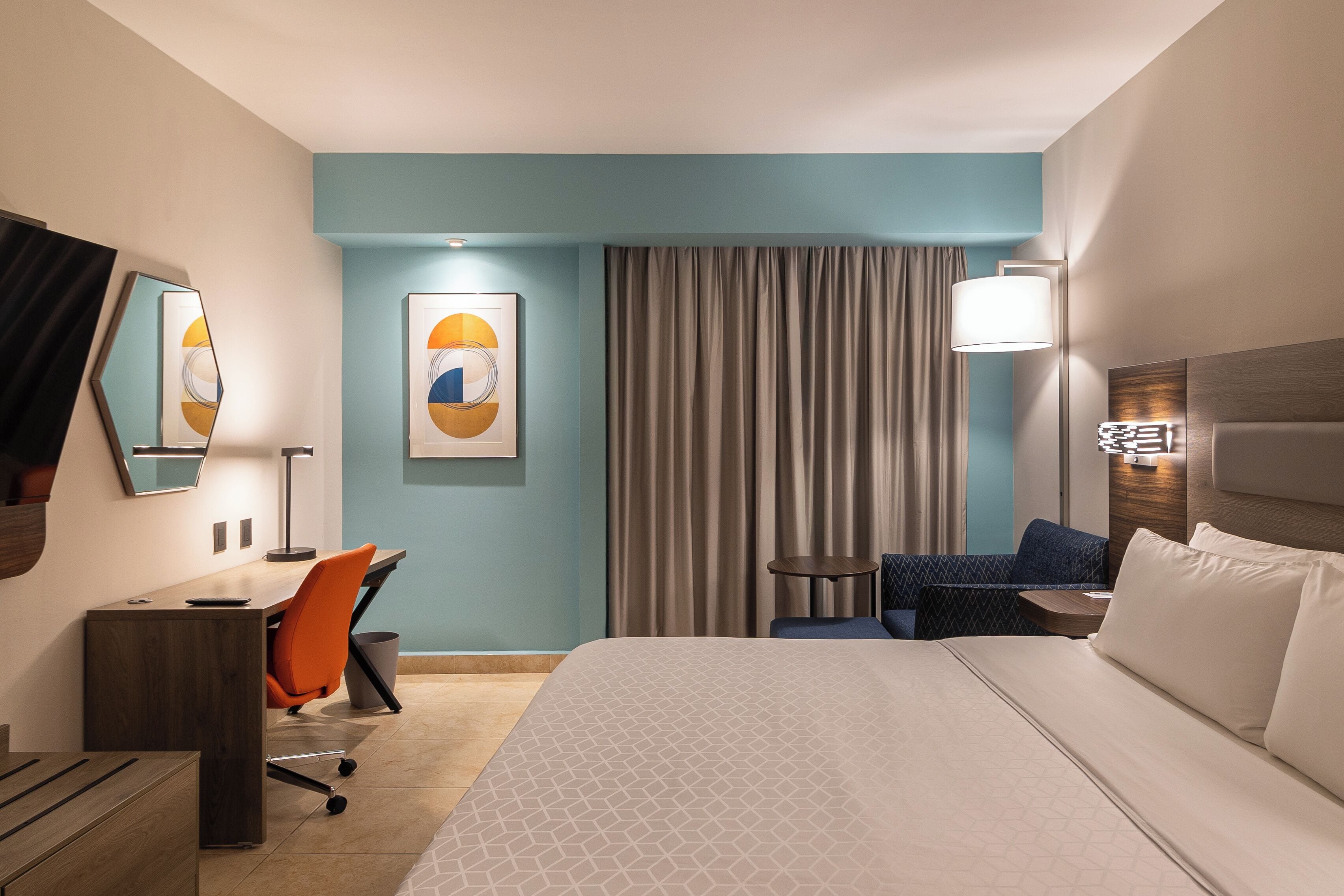 Ett hotellrum med en stor säng, ett skrivbord med en orange stol, ett litet bord, en lampa och ett stort fönster med gardiner.