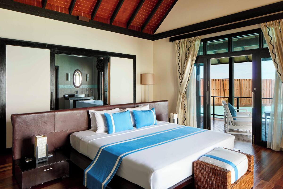 A hotel room with a large bed, a nightstand, a mirror, a window with curtains, and a view of a wooden deck.