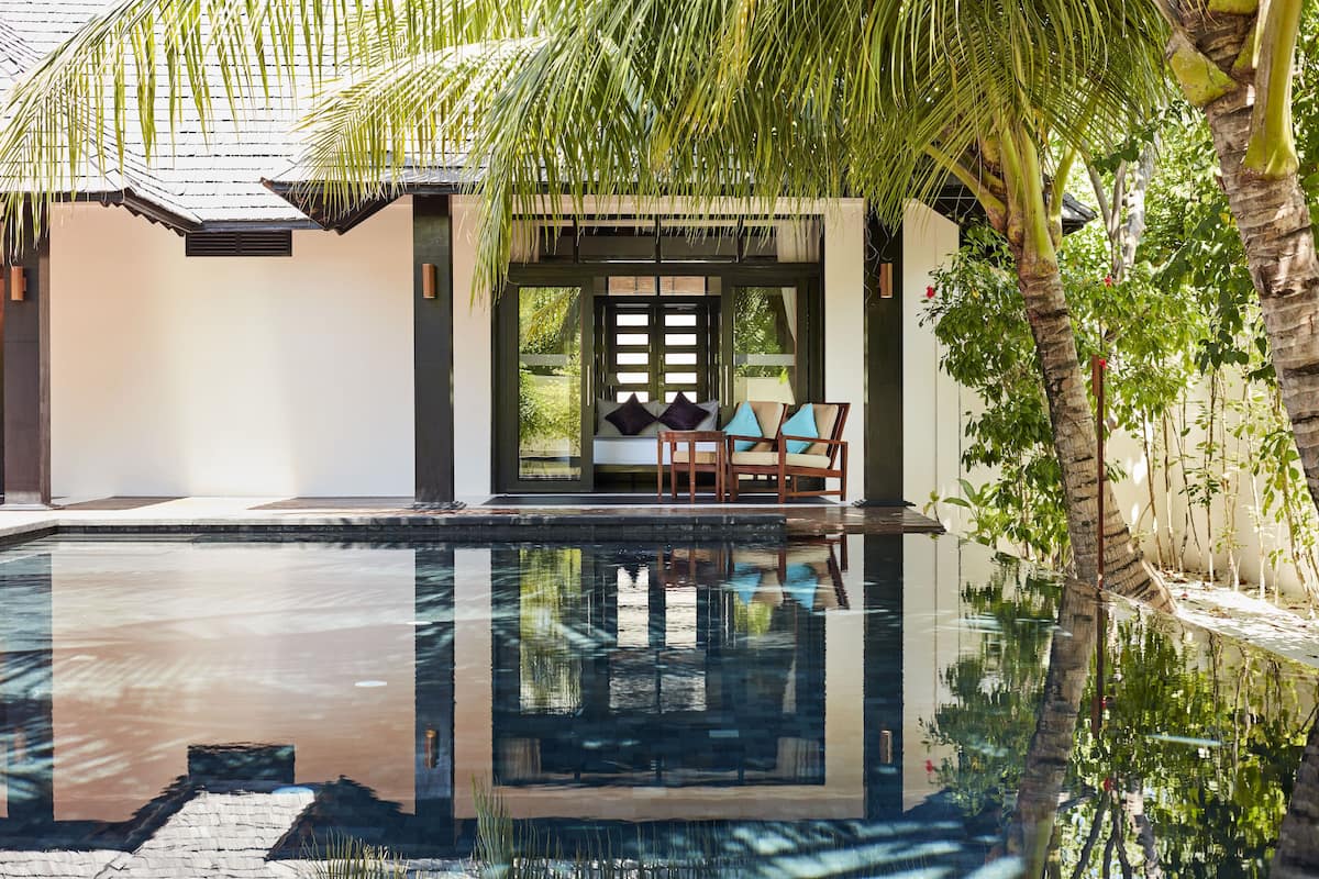 A serene pool area with a clear reflection, a seating area with cushions, and a palm tree.