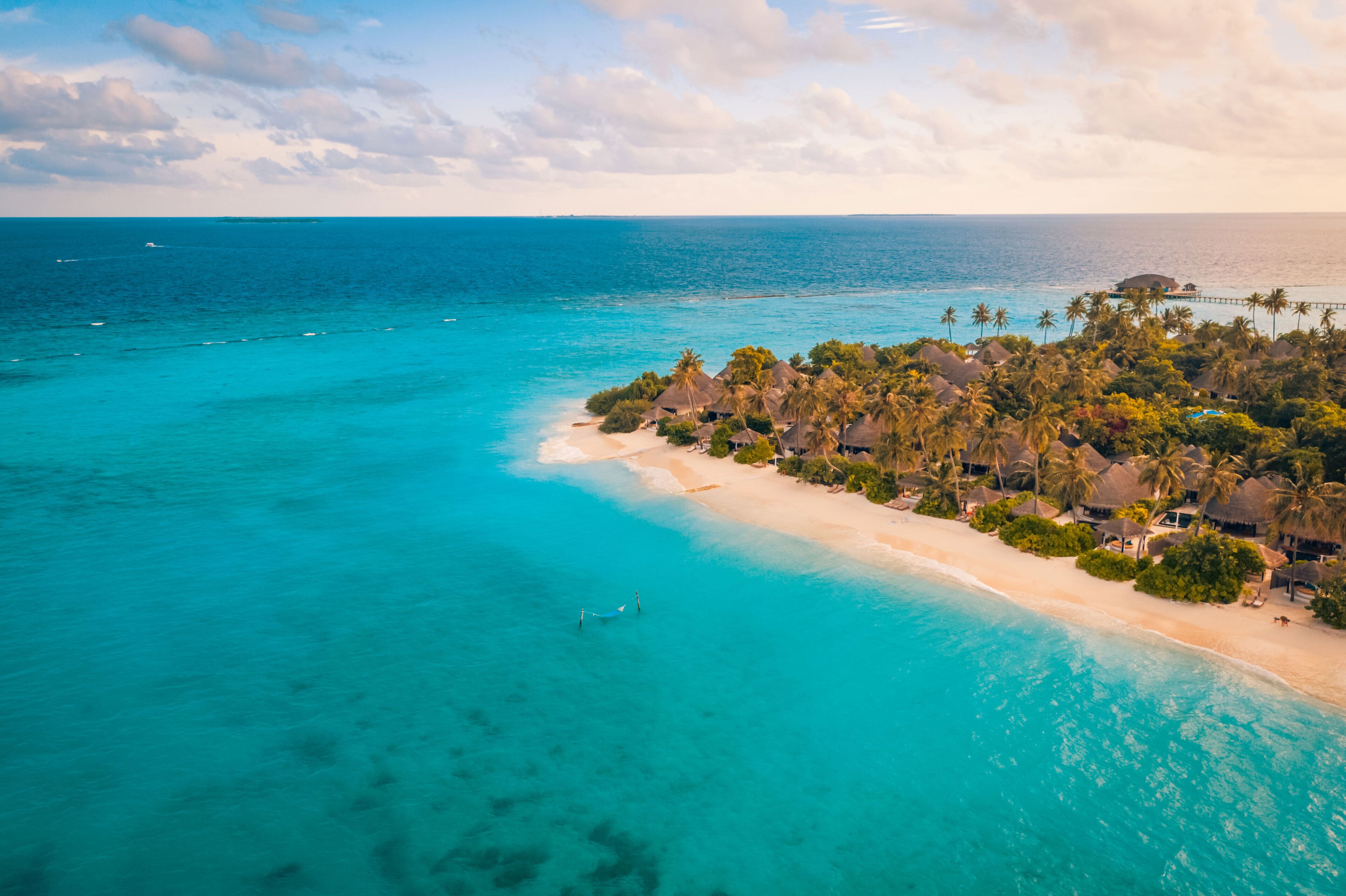 An aerial view of a tropical beach resort with clear turquoise waters, a sandy beach, and palm trees.