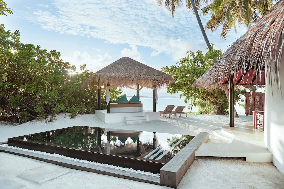A pool area with a thatched roof structure, lounge chairs, and a view of the sea.