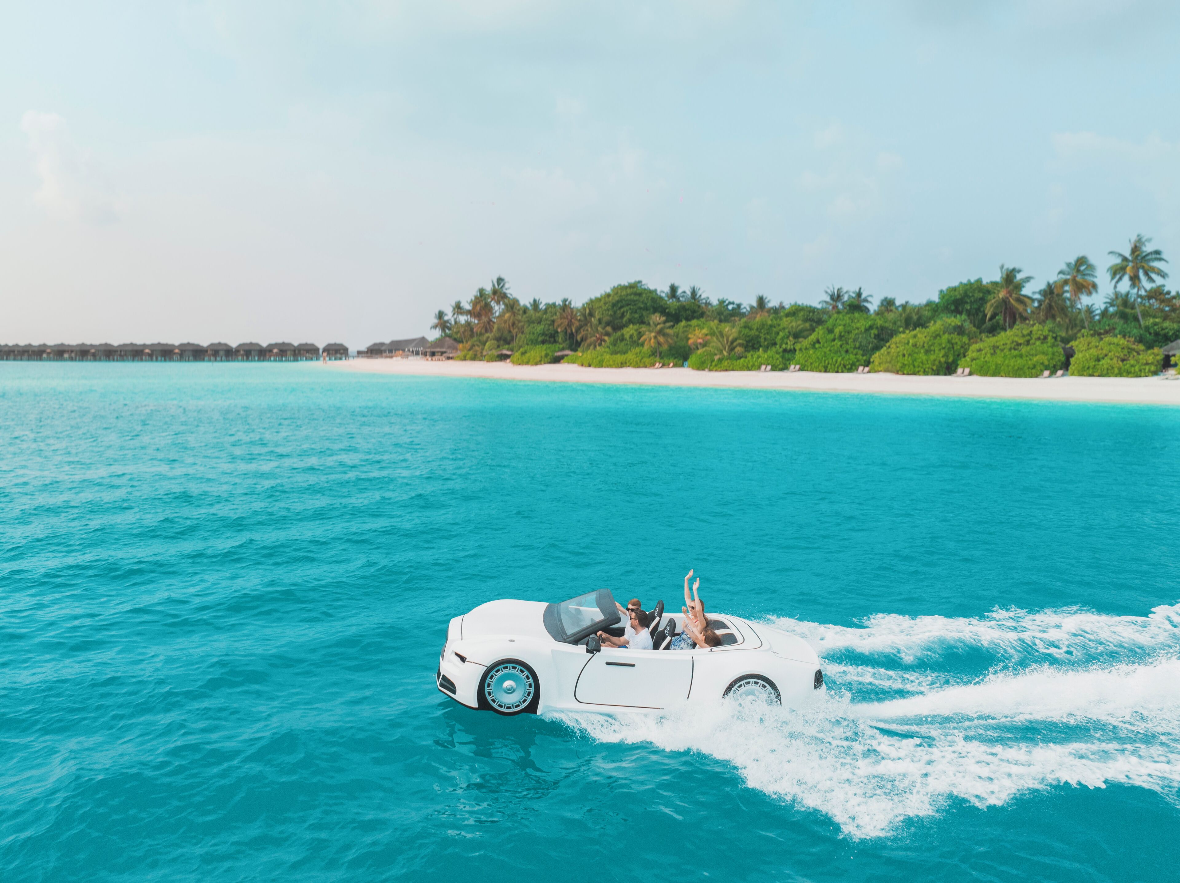 Un coche descapotable blanco circulando sobre aguas turquesas, con una playa y palmeras al fondo.
