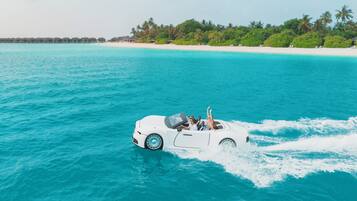 Un coche descapotable blanco circulando sobre aguas turquesas, con una playa y palmeras al fondo.