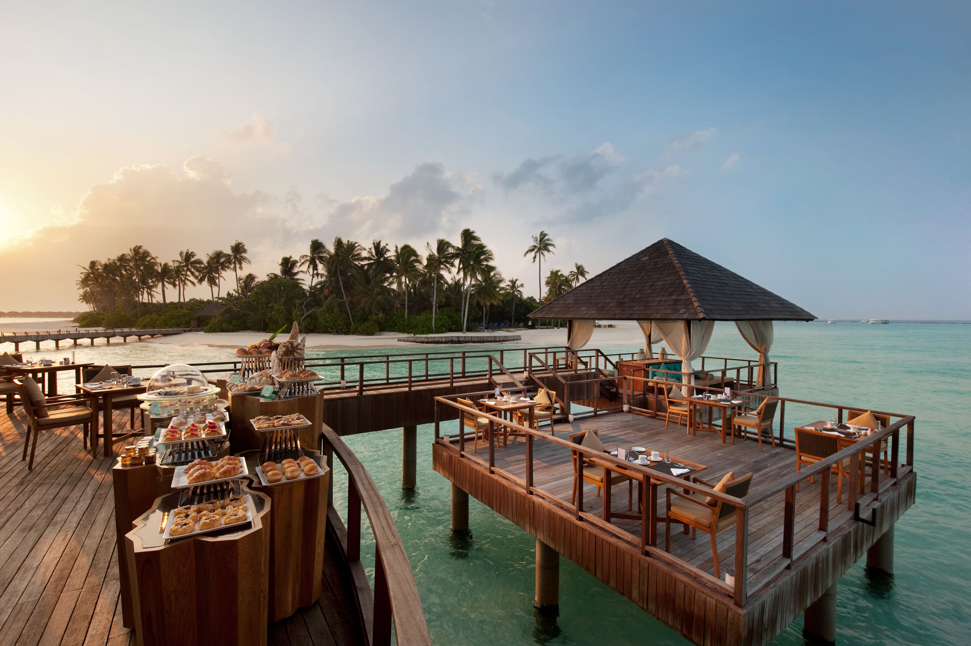 Una terraza de madera con mesas y sillas de comedor, una zona de buffet y un pabellón con vistas a una playa con palmeras y agua azul cristalina.