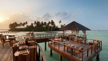 Una terraza de madera con mesas y sillas de comedor, una zona de buffet y un pabellón con vistas a una playa con palmeras y agua azul cristalina.