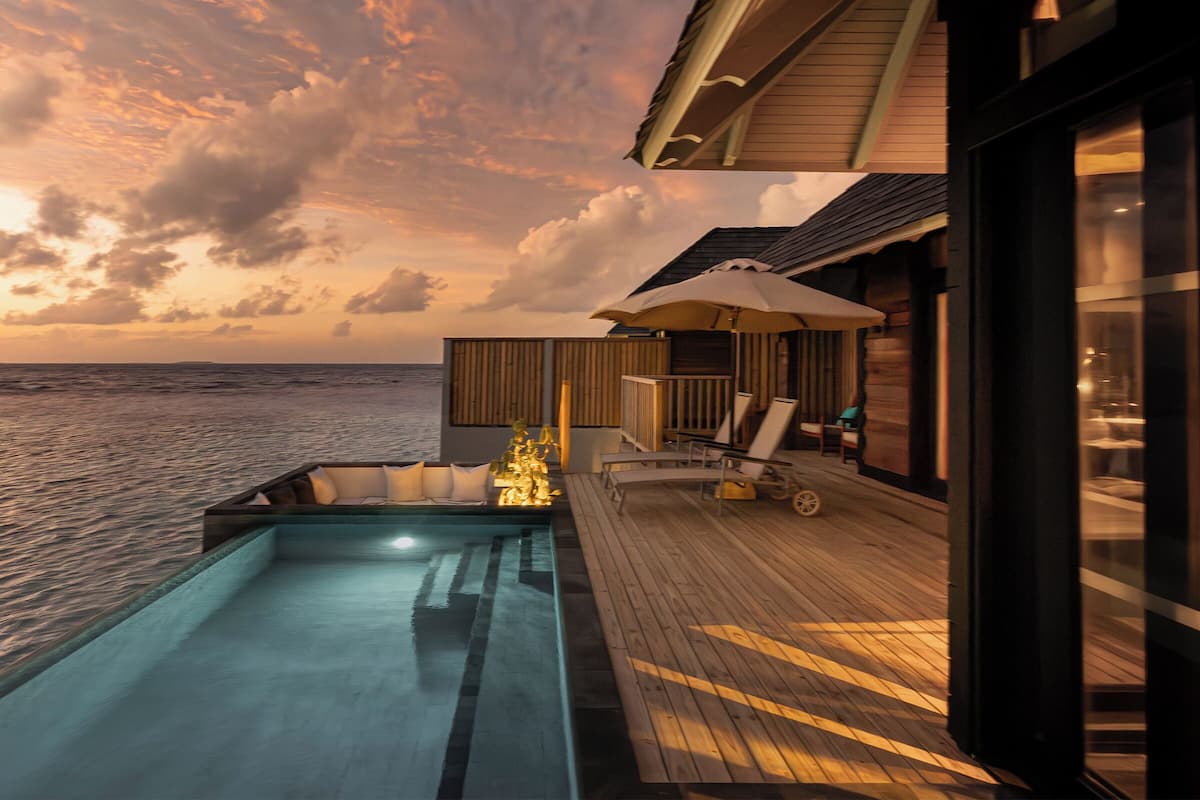 A poolside area with a view of the ocean at sunset, wooden deck, lounge chairs, and a small structure with a roof.