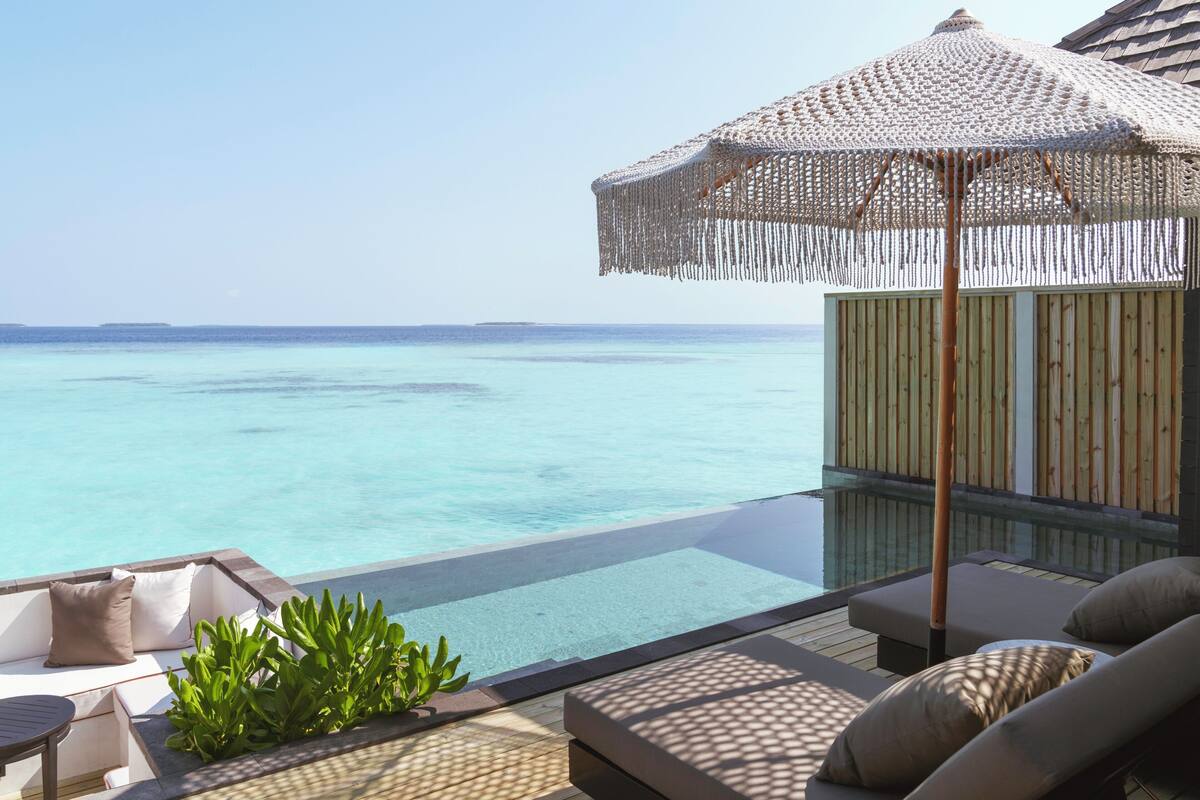 An infinity pool with clear blue water, a lounge area with cushions, and a straw umbrella.