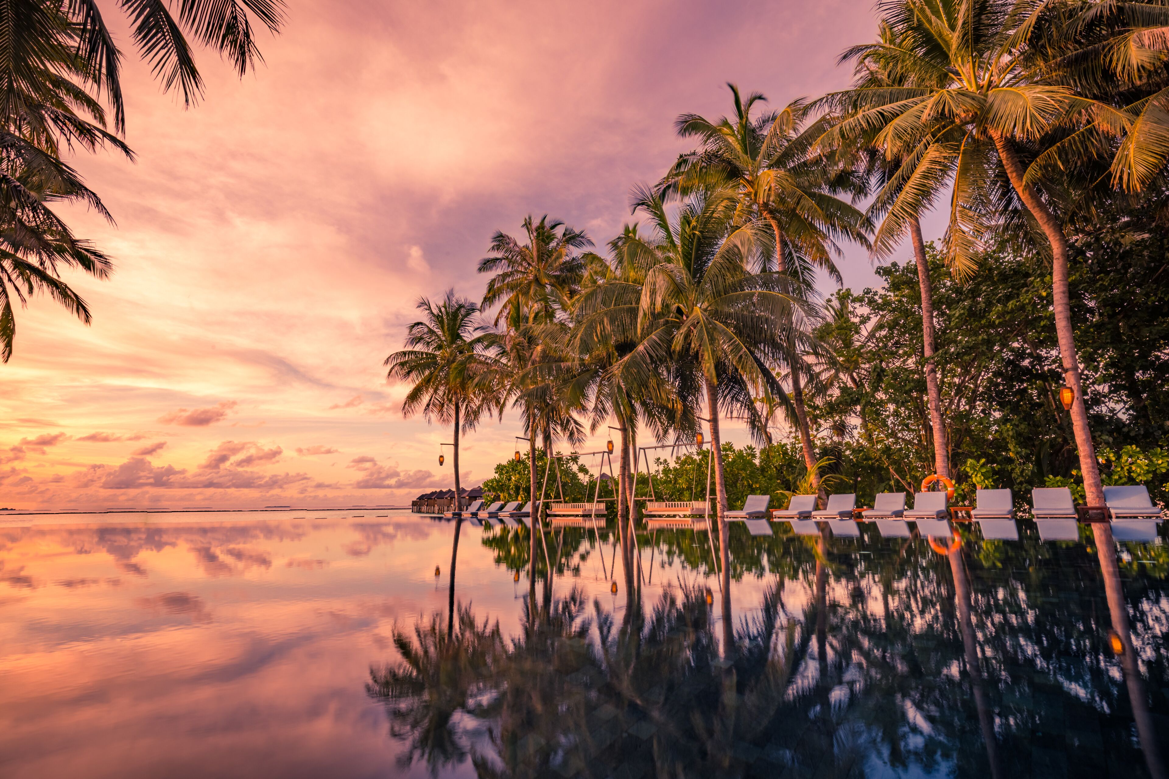 Una playa tranquila con palmeras y una piscina infinita que refleja el cielo.