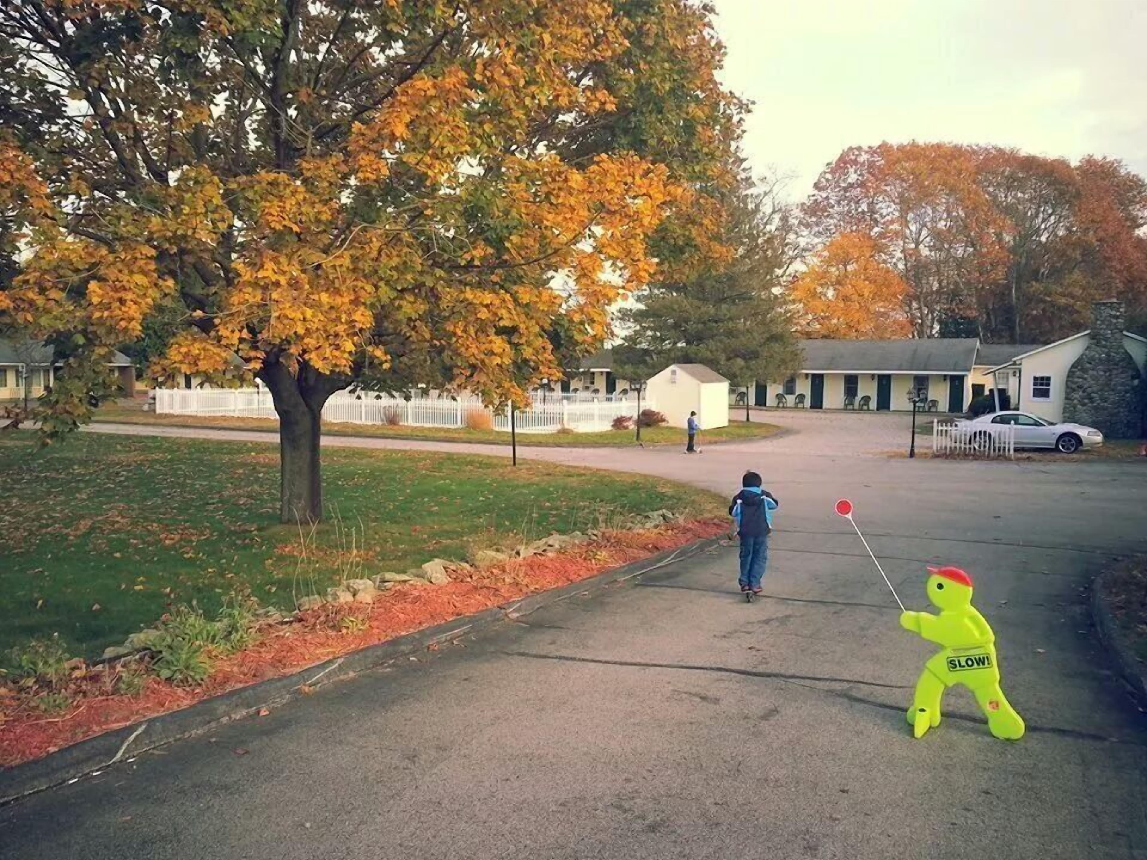 A residential street with houses, a tree with autumn leaves, and a child playing with a toy.