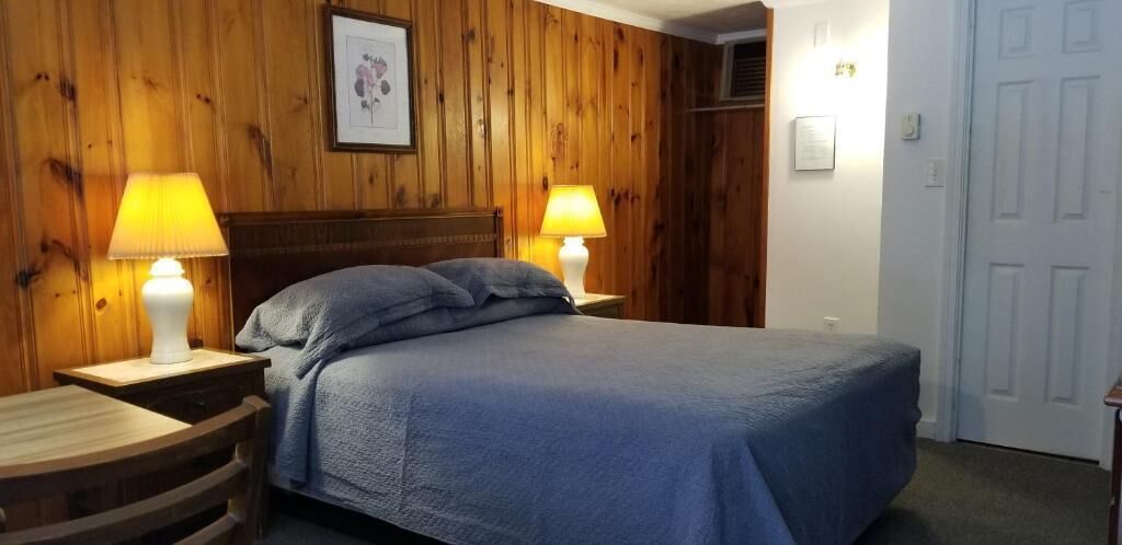 Een slaapkamer met een houten wandpaneel, een bed met een blauwe sprei, twee bedlampen en een ronde houten stoel.
