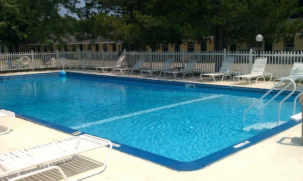Piscina rectangular con agua azul cristalina, rodeada por una cerca blanca y múltiples sillones de descanso.
