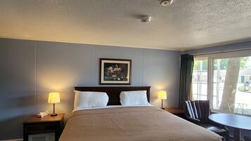 A hotel room with a bed, two bedside lamps, a painting on the wall, a window with a view of the outside, and a small table with a chair.