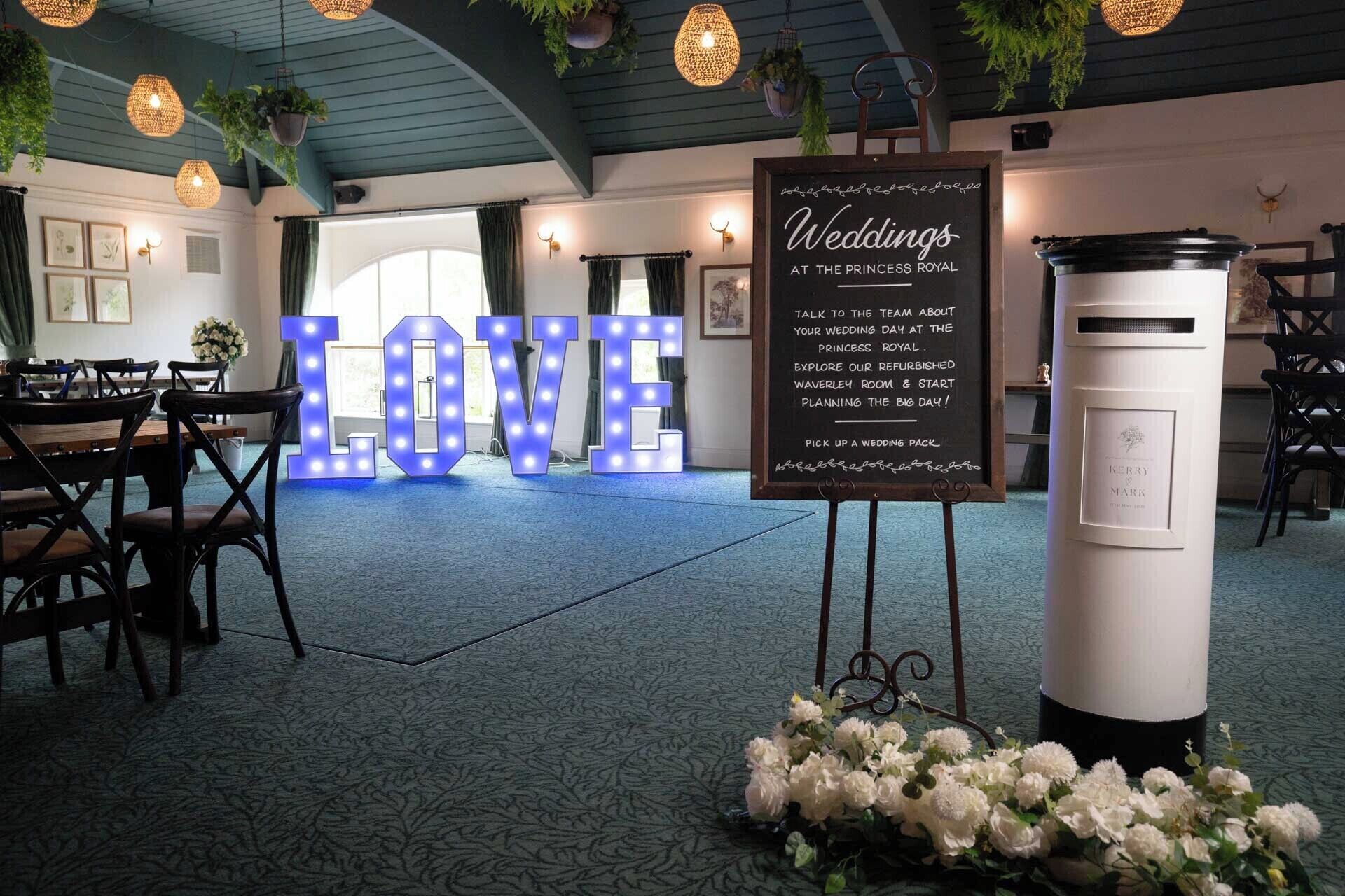 A wedding venue with a large illuminated 'LOVE' sign, a chalkboard with wedding details, and a white column with a plaque.