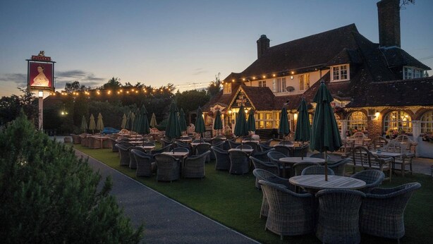 An outdoor seating area with wicker chairs and tables, a building with a sign reading "The Princess Royal," and string lights in the background. - The Princess Royal (Farnham)