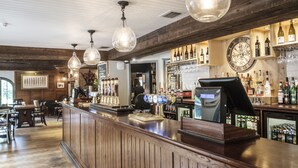 A bar area with wooden paneling, a counter with a cash register, and a variety of bottles and glasses on shelves. - The Princess Royal (Farnham)