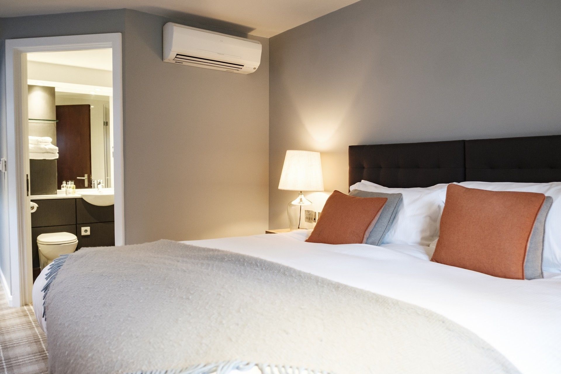 A hotel room with a bed, a nightstand, a lamp, an air conditioning unit, and a bathroom visible through an open door.