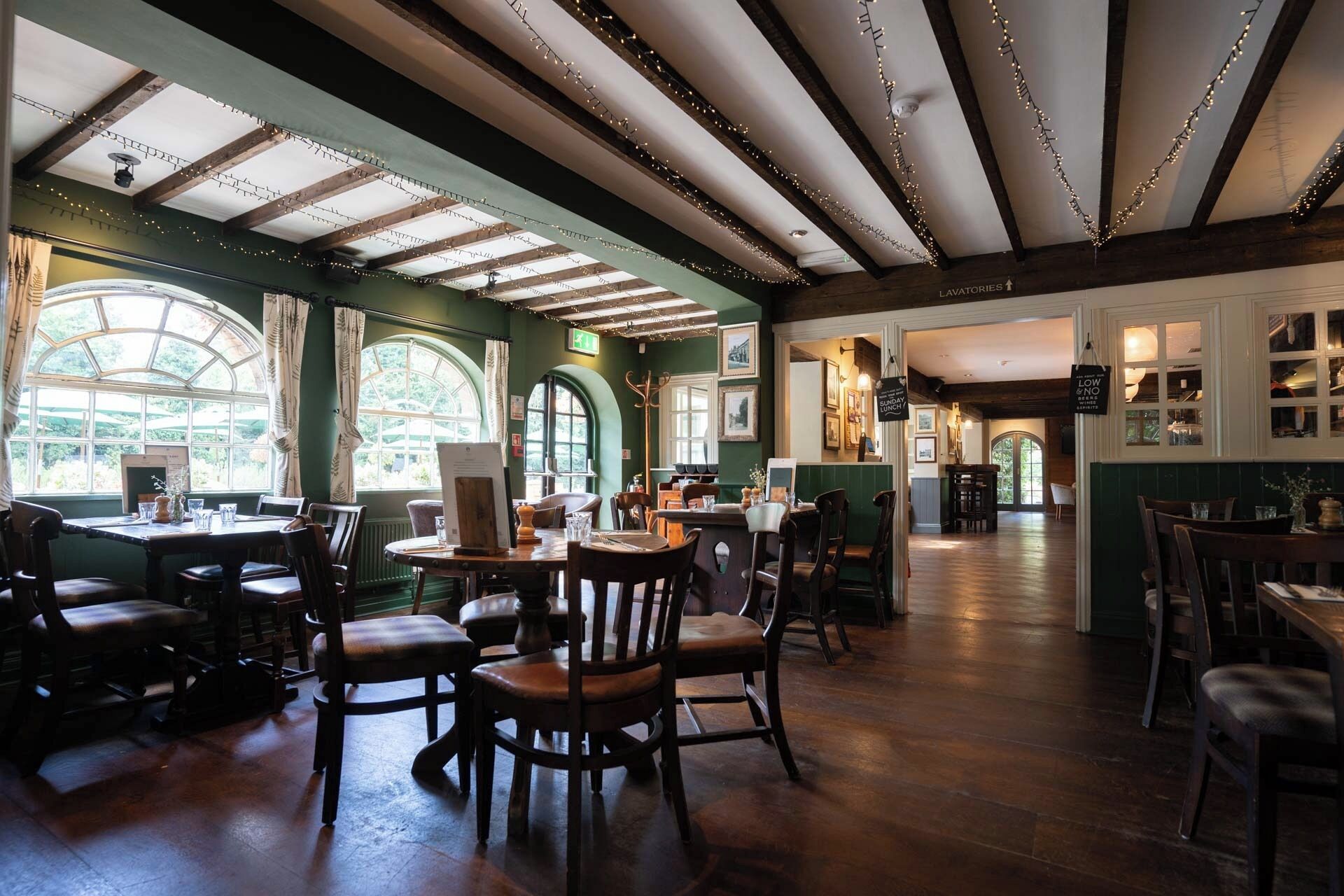 Un ristorante con tavoli e sedie di legno, un soffitto alto con travi a vista e ampie finestre che lasciano entrare la luce naturale.