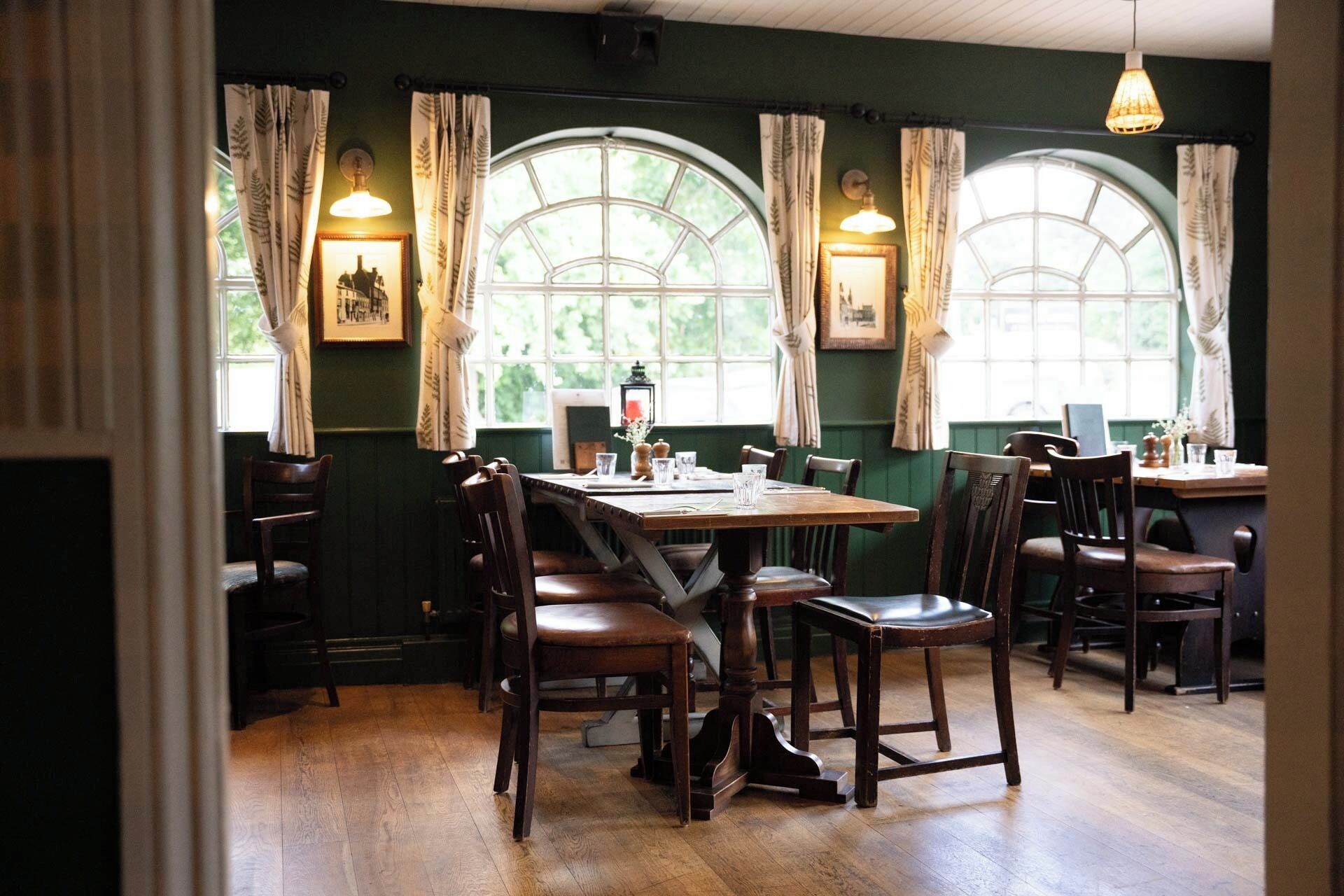 Un espace repas avec des tables et des chaises en bois, de grandes fenêtres en arc surmontées de rideaux et des lampes fixées au mur.