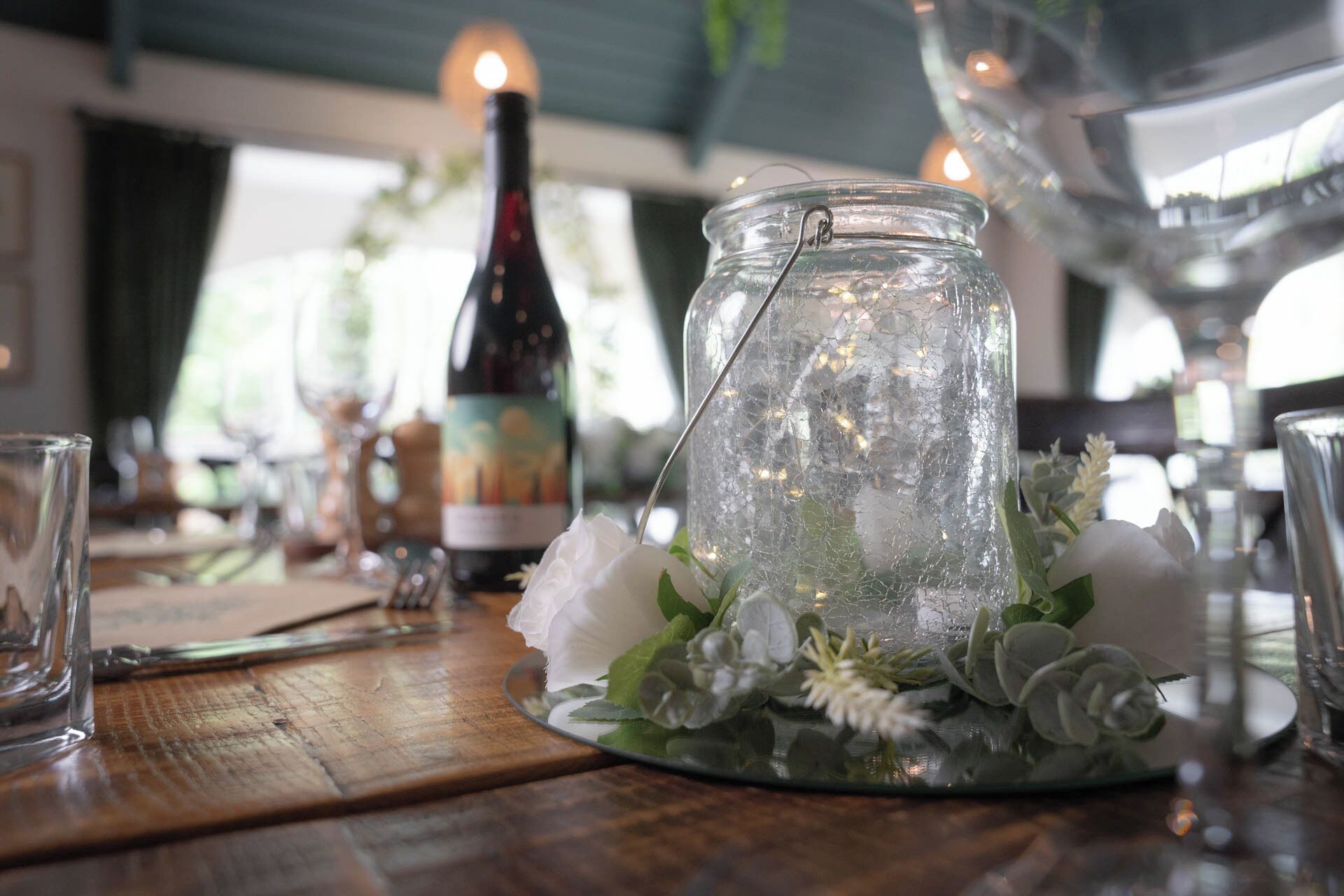 Una mesa con un tarro de vidrio que contiene flores y follaje, una botella de vino y copas.