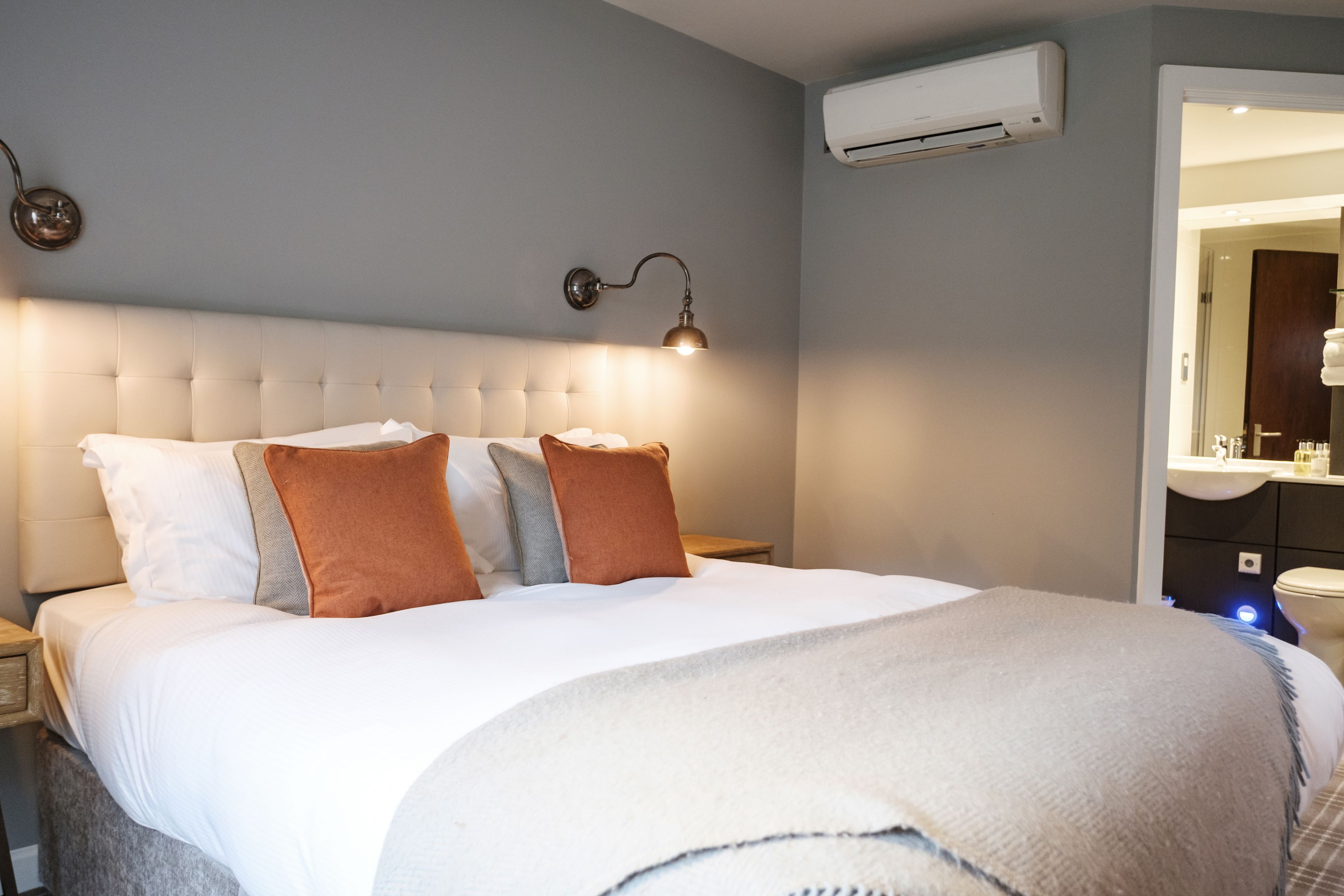 A neatly made bed with a white duvet and several pillows, a wall-mounted air conditioner, and a bedside lamp.