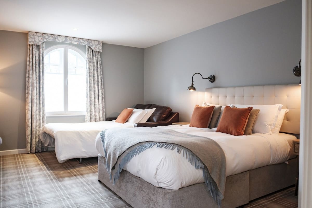 Ein Schlafzimmer mit einem großen Bett, einem kleineren Bett, einem Nachttisch, einer Lampe und einem Fenster mit Vorhängen.