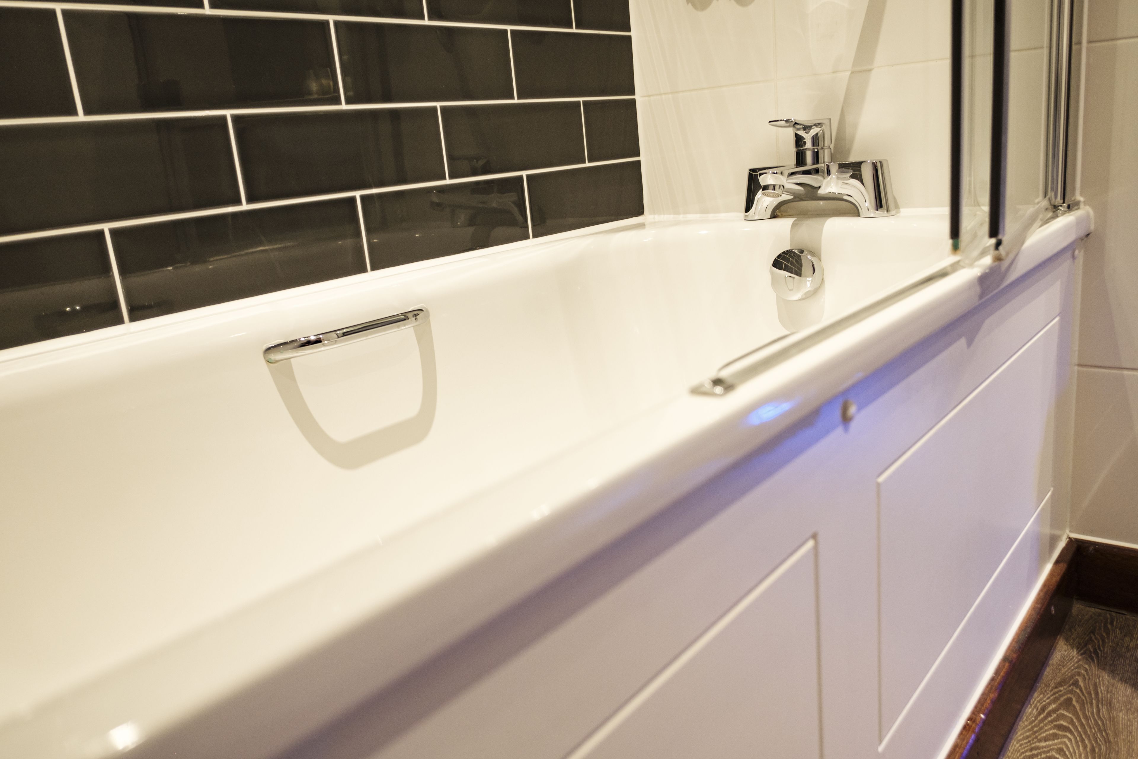 A white bathtub with a chrome faucet and a handrail, set against a black tiled wall.