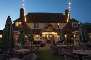 A traditional pub with a sign reading "The Princess Royal". It features outdoor seating with wicker chairs and tables, and is illuminated by string lights.