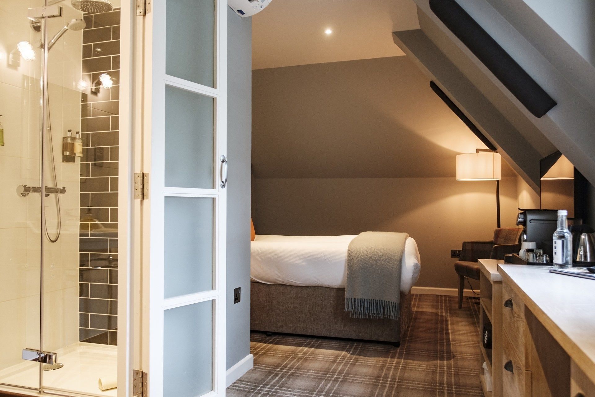 Habitación de hotel con ducha, cama, mesita de noche, lámpara y cafetera.