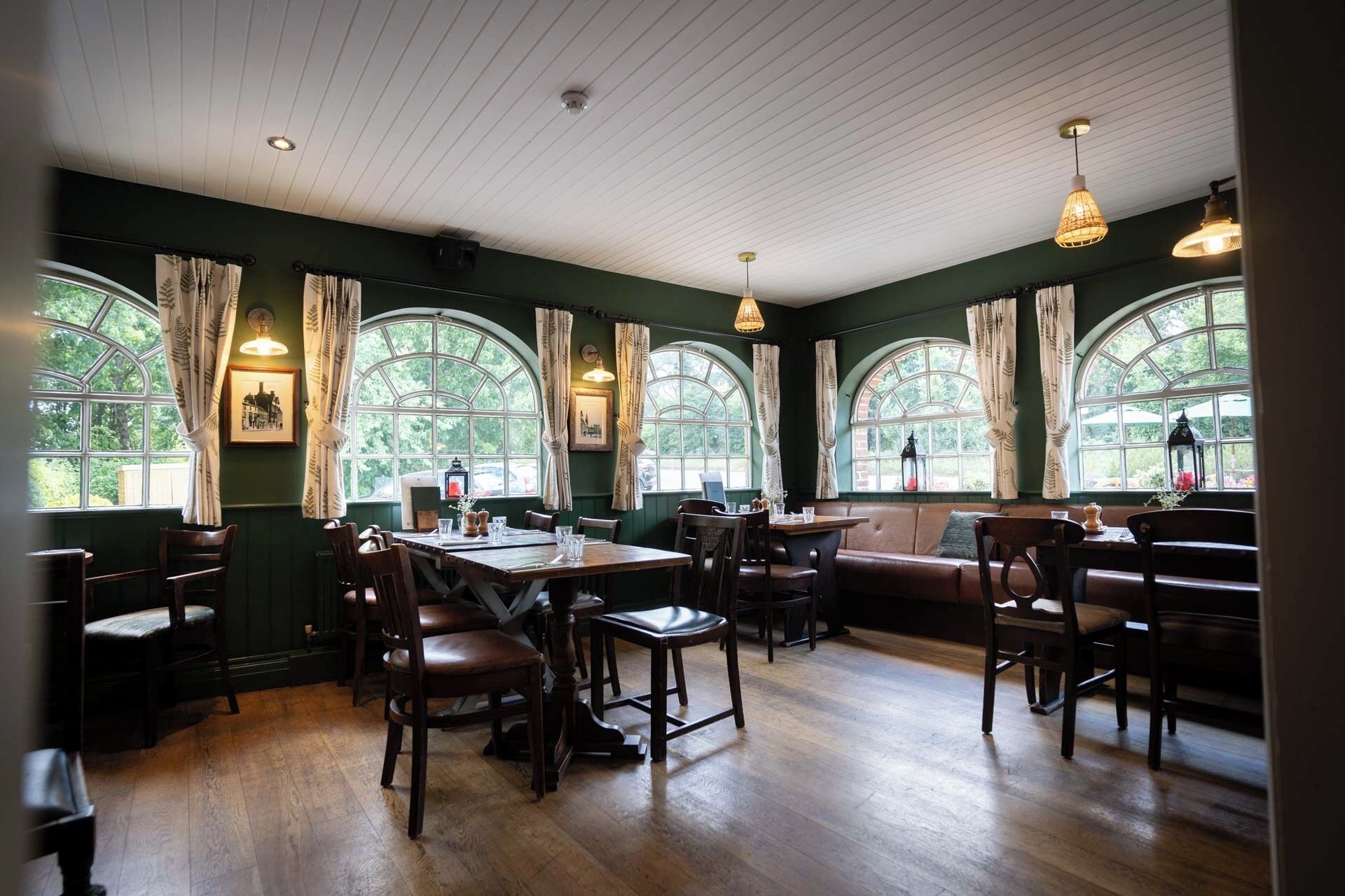 A restaurant with wooden tables and chairs, large arched windows, and hanging pendant lights.