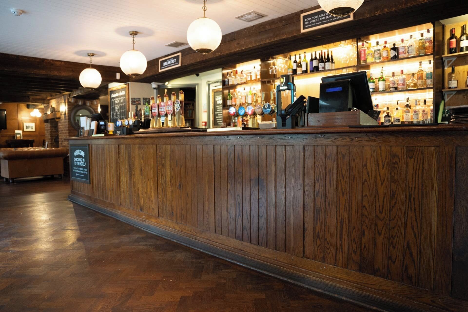 A bar area with wooden paneling, a counter with a sign, and a variety of bottles on shelves.